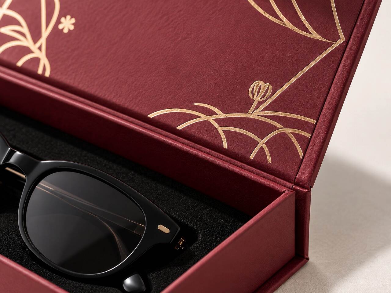Close-up of burgundy specialty paper, gold foil botanical artwork, wrapped rigid-box edges and black eyewear insert Close-up of burgundy specialty paper, gold foil botanical artwork, wrapped rigid-box edges and black eyewear insert